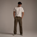 A man stands against a plain background, wearing a white t-shirt and T7C’s The Liam Military Pant in Olive Drab with black sneakers. His hands are in his pockets and he looks down to his left.