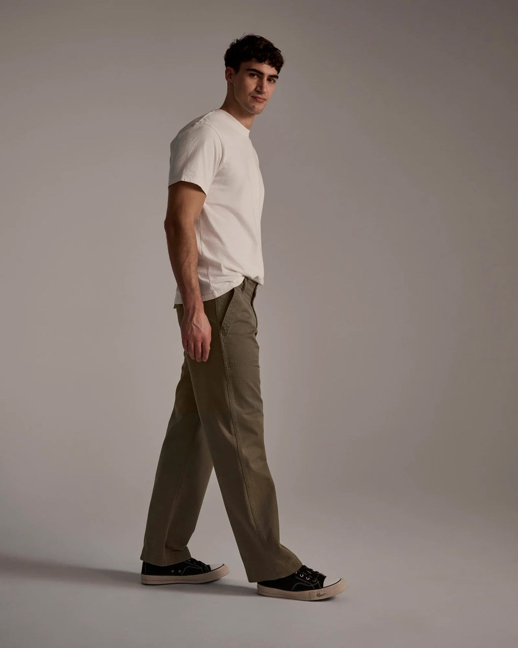 A man wears T7Cs The Liam Military Pant in Olive Drab, paired with a white t-shirt and black sneakers, walking in a studio with a neutral background and glancing over his shoulder.