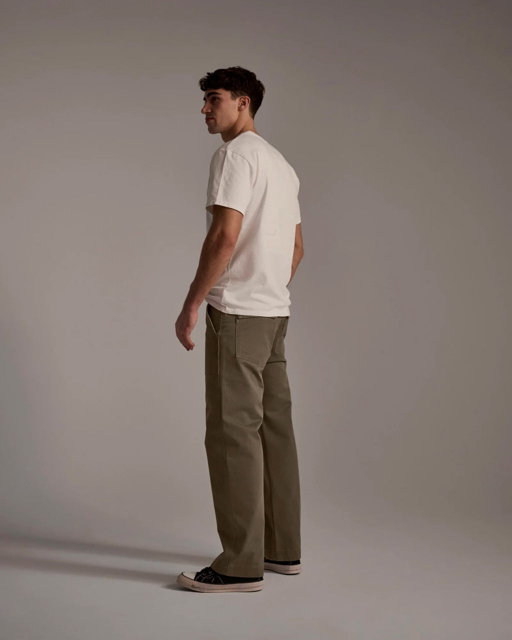 A man stands in a studio with a neutral background, wearing a white t-shirt and T7C’s The Liam Military Pant in Olive Drab. He completes the look with black sneakers, turned slightly to the side and looking away from the camera.