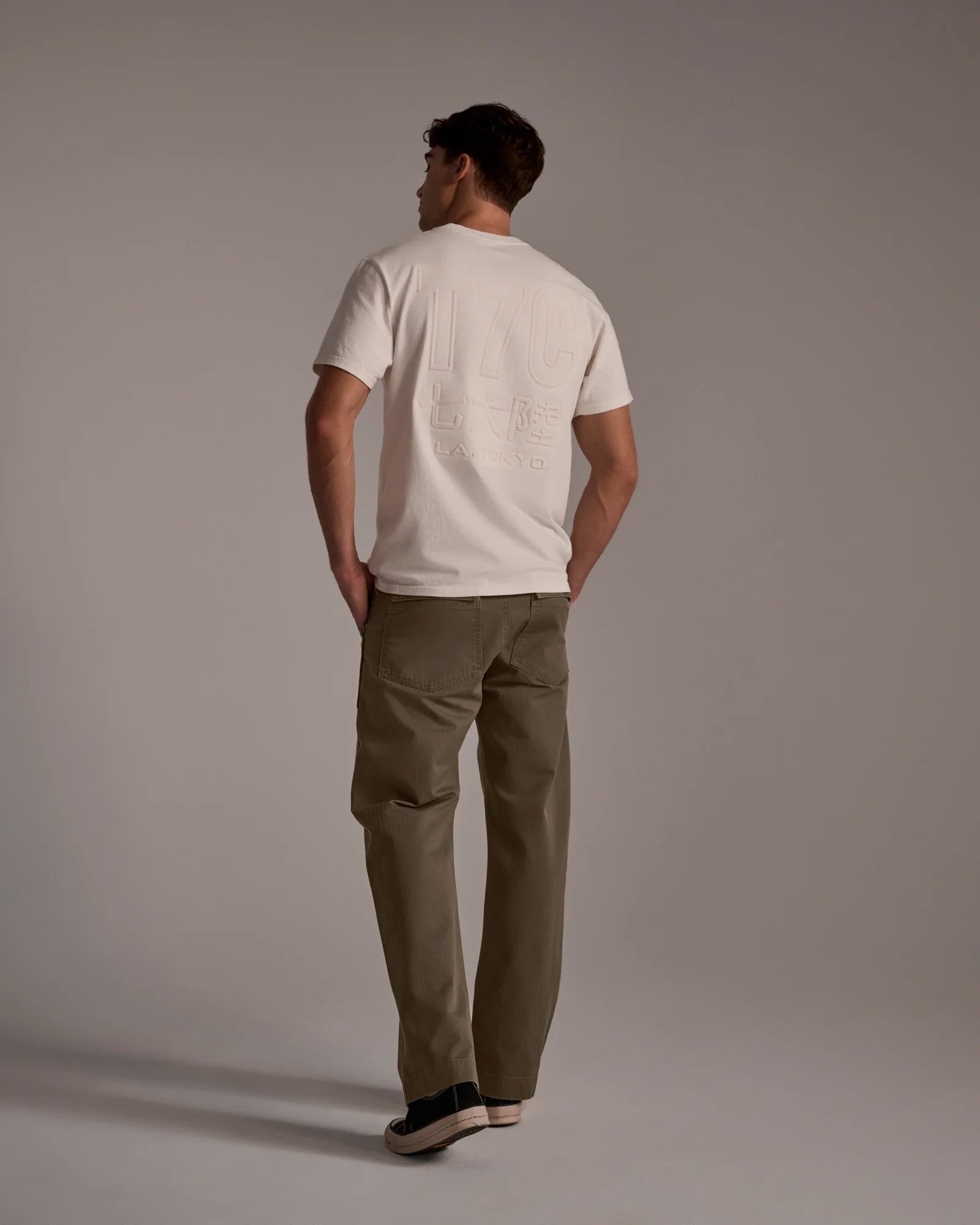 A man faces away in a studio, wearing a white T-shirt with embossed back graphics, The Liam Military Pant in Olive Drab by T7C, and black sneakers. The background is plain and neutral.