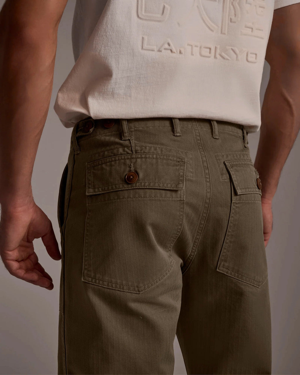 A person wears the T7C Liam Military Pant in Olive Drab, featuring relaxed fit and buttoned back pockets, paired with a white T-shirt with “L.A. TOKYO” text. The photo highlights the back view from waist to mid-thigh.