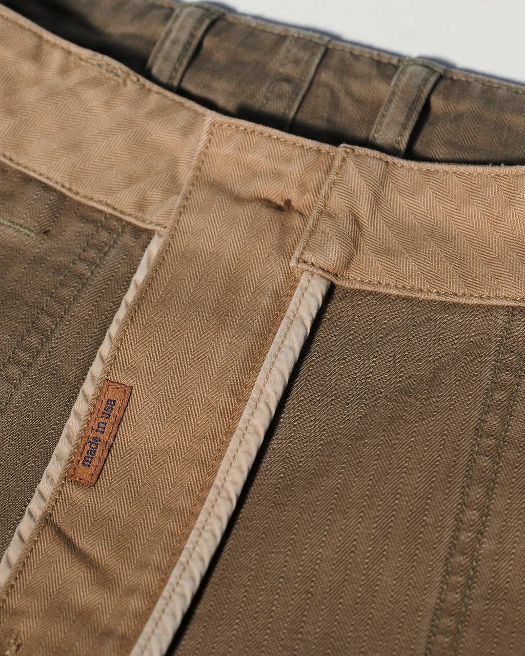 Close-up of the waistband and inner stitching of The Liam Military Pant in Olive Drab by T7C, crafted from cotton herringbone fabric with detailed patterns, belt loops, and a small brown label featuring blue Sun House text.