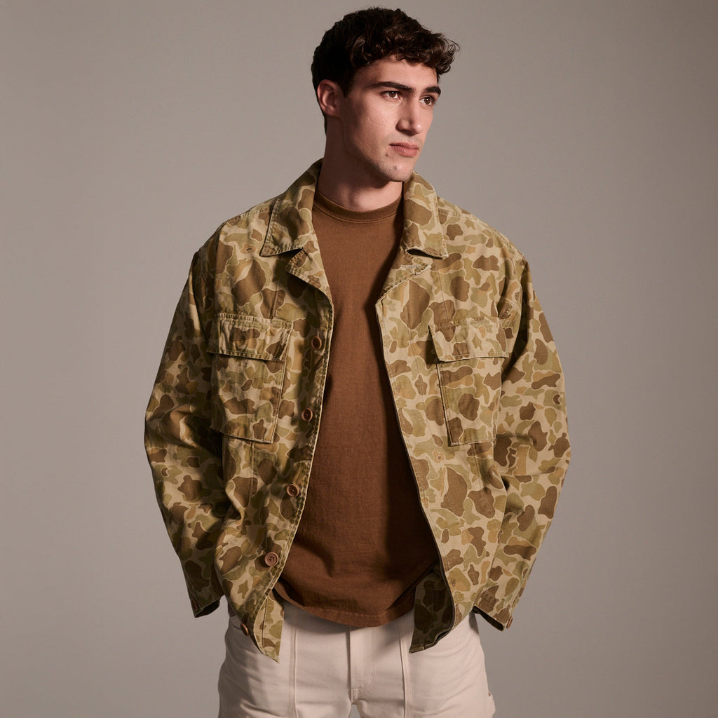 A man stands against a plain background wearing The Ryan Jacket in Camouflage by T7C The Seven Continents over a brown shirt and light pants, looking slightly to the side with a neutral expression.