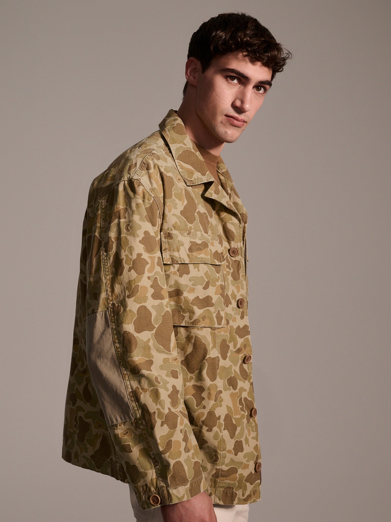 A man with short dark hair wears the T7C The Seven Continents Ryan Jacket in Camouflage, a relaxed-fit overshirt featuring light brown and green camo, standing against a plain gray background.