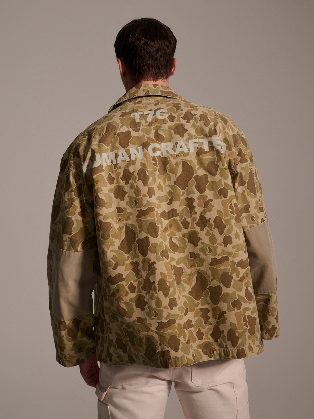 A man faces away from the camera, wearing T7C The Seven Continents The Ryan Jacket in Camouflage with HUMAN CRAFTS and 1767 in white on the back, paired with light-colored pants.