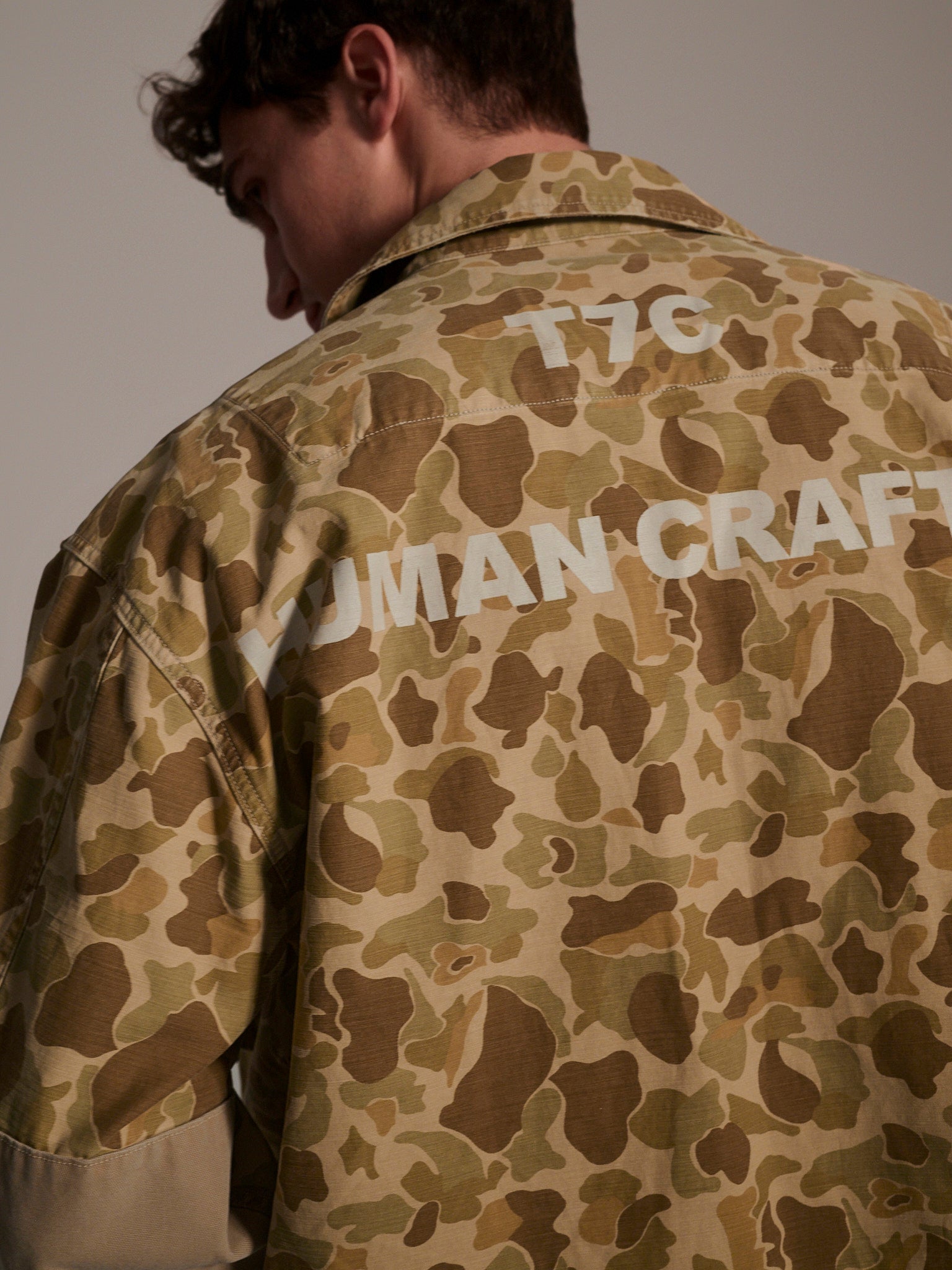 A person shown from behind and slightly to the side wears The Ryan Jacket in Camouflage by T7C The Seven Continents, a relaxed-fit beige and brown overshirt with “HUMAN CRAFT” and “F/7C” in white on the back.