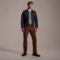 A man stands in a studio wearing a blue denim jacket, gray shirt, T7C The Seven Continents Sid Pant in Hide Raw, and black boots against a plain neutral background.
