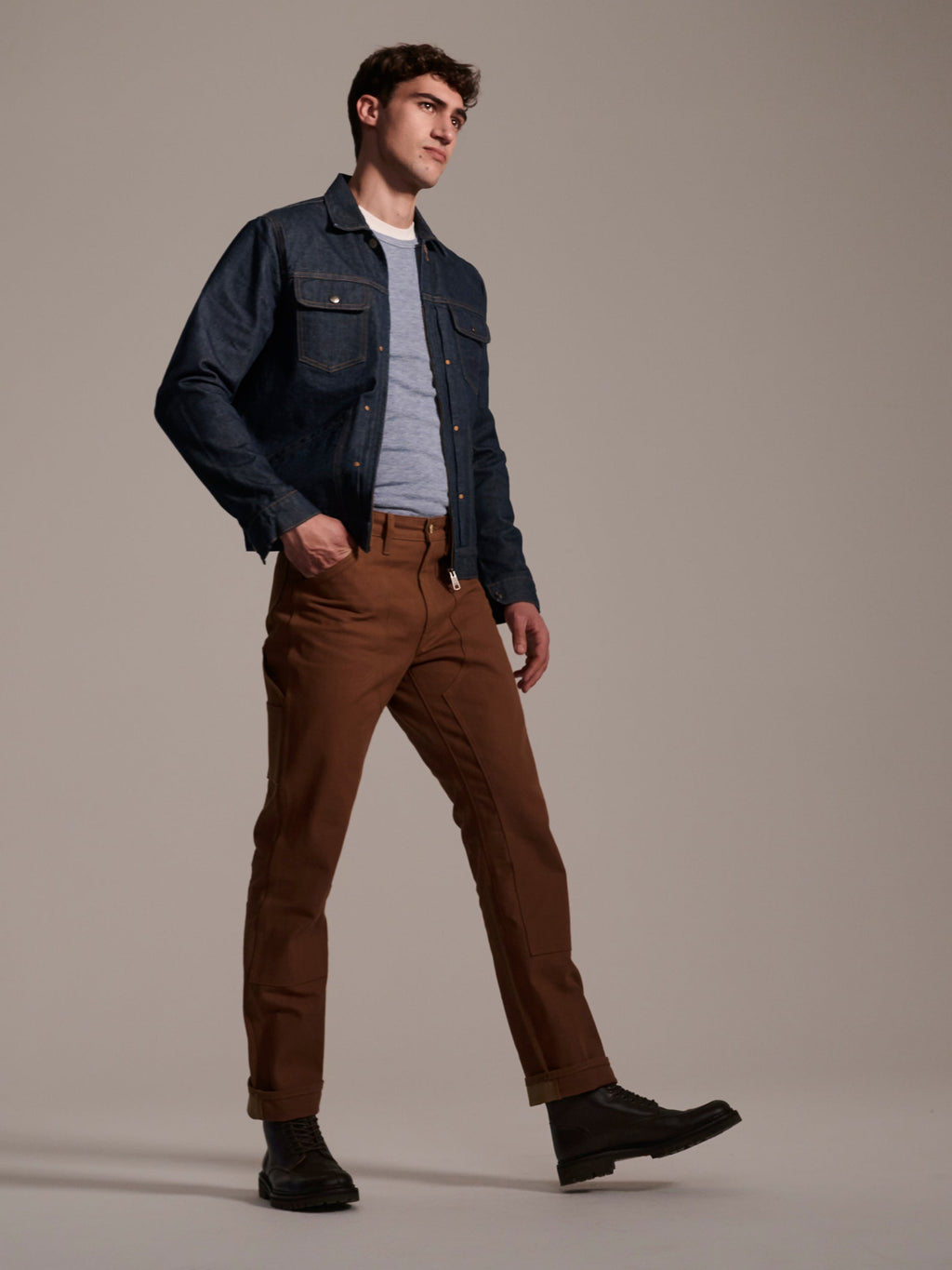 A man in a denim jacket, gray tee, and T7C The Seven Continents The Sid Pant in Hide Raw strides in black boots against a plain beige background.
