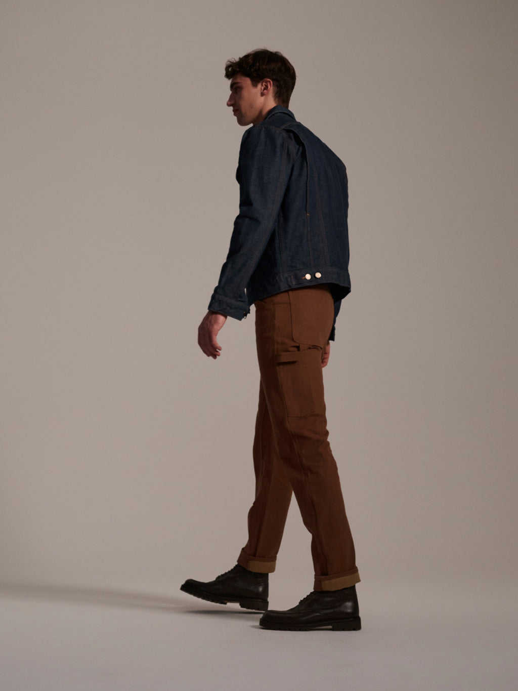 A man walks left against a light background, wearing a blue denim jacket, black shoes, and T7C The Seven Continents’ The Sid Pant in Hide Raw for a classic workwear look.
