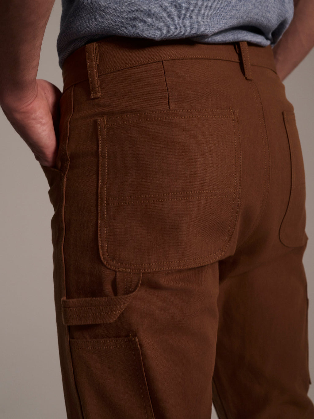 A close-up of someone wearing T7C The Seven Continents’ The Sid Pant in Hide Raw, featuring large back pockets and a hand in the side pocket, paired with a gray shirt—perfect for functional workwear.