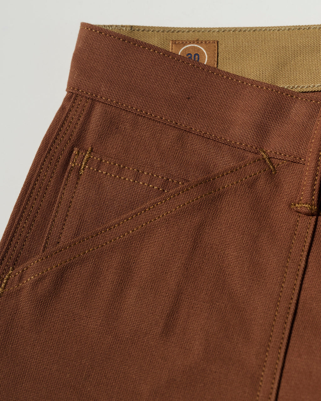 Close-up of T7C The Seven Continents The Sid Pant in Hide Raw—slim-straight brown pants featuring detailed stitching, front pocket, belt loop, and a small tan 30 label at the waistband for classic workwear style with refined utility.