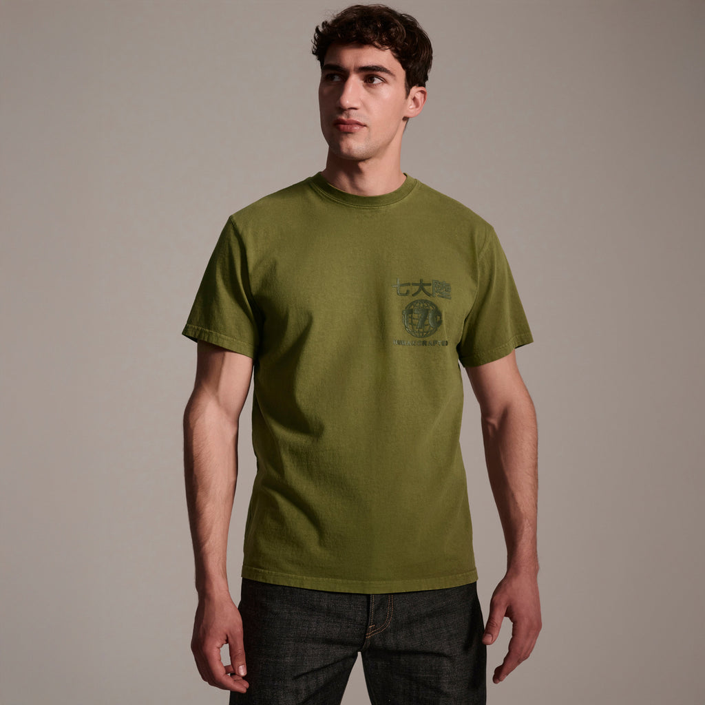 A man with short brown hair wears The Timo Tee in Olive by T7C The Seven Continents, featuring a puff screen printed logo and chest text, paired with dark jeans against a plain beige background.