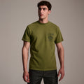 A man with short brown hair wears The Timo Tee in Olive by T7C The Seven Continents, featuring a puff screen printed logo and chest text, paired with dark jeans against a plain beige background.