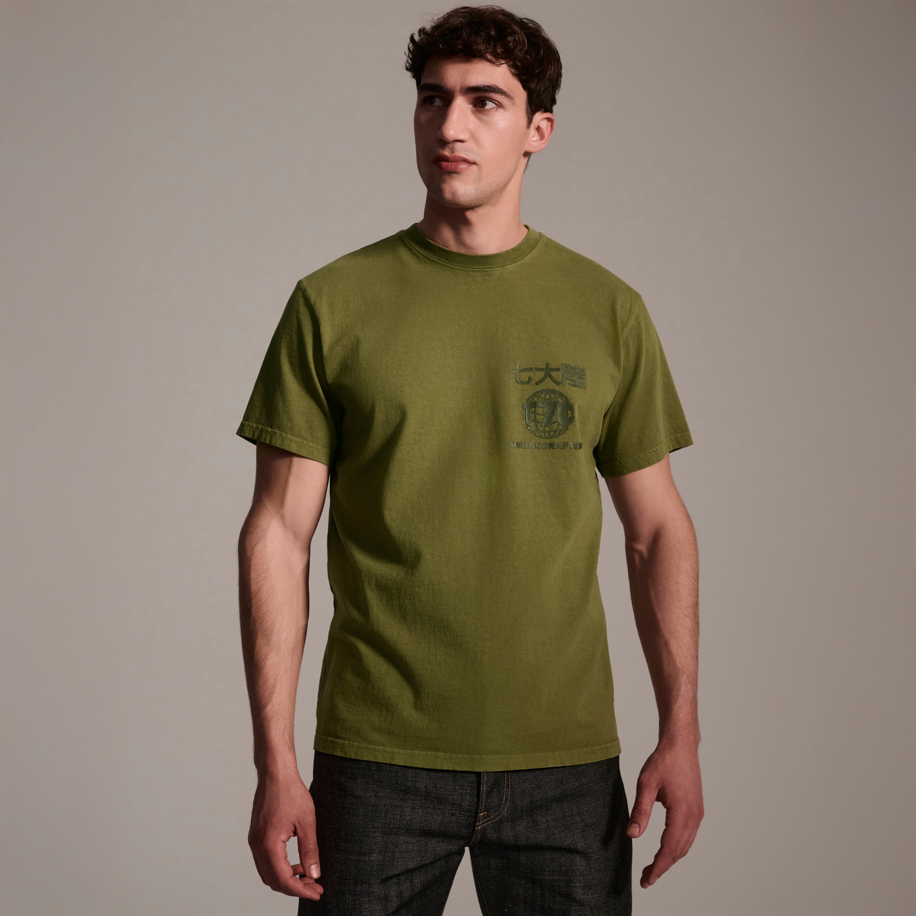 A man with short brown hair wears The Timo Tee in Olive by T7C The Seven Continents, featuring a puff screen printed logo and chest text, paired with dark jeans against a plain beige background.