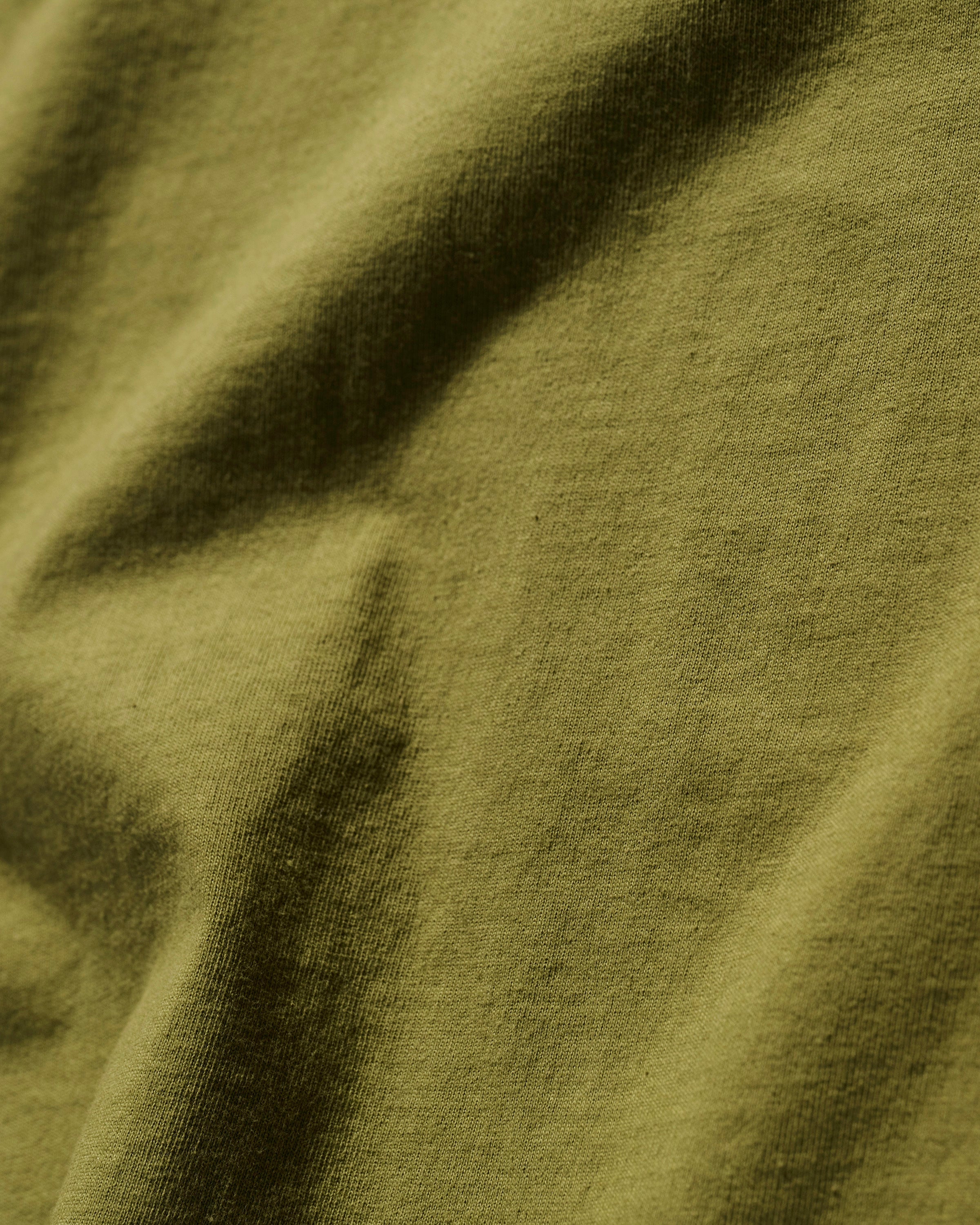 Close-up of The Timo Tee in Olive by T7C The Seven Continents, featuring ultra-soft fabric with gentle folds and a puff screen printed logo. Soft shadows and highlights accentuate the textured surface.