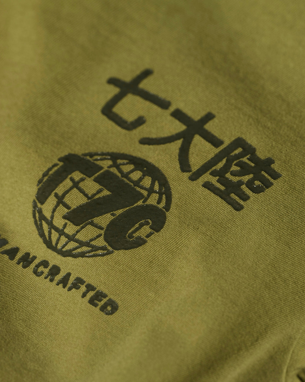 Close-up of The Timo Tee in Olive by T7C The Seven Continents, featuring extra soft fabric, Japanese and English text, a globe with T7C, “HANDCRAFTED” detail, and a puff screen printed logo.