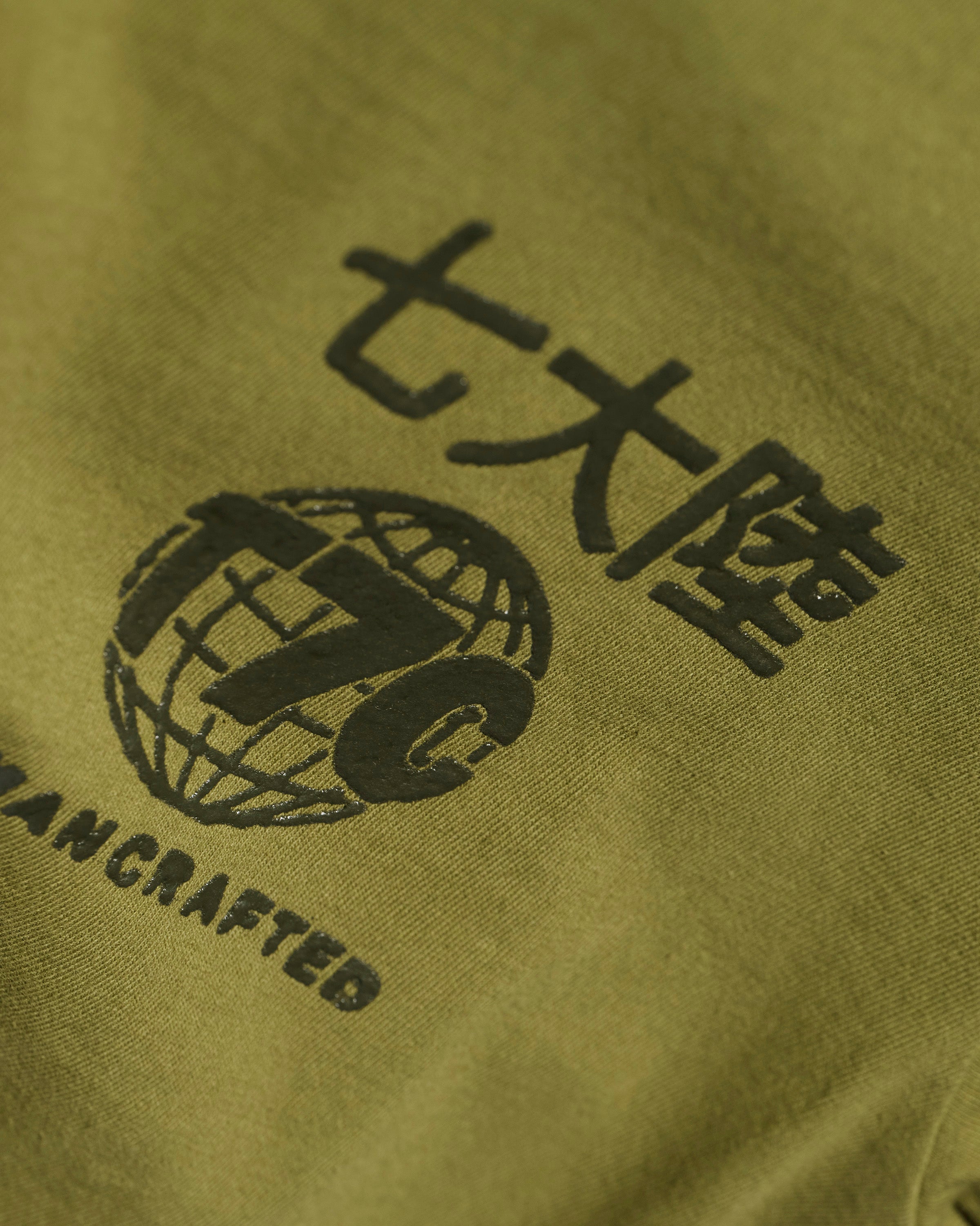 Close-up of The Timo Tee in Olive by T7C The Seven Continents, featuring extra soft fabric, Japanese and English text, a globe with T7C, “HANDCRAFTED” detail, and a puff screen printed logo.