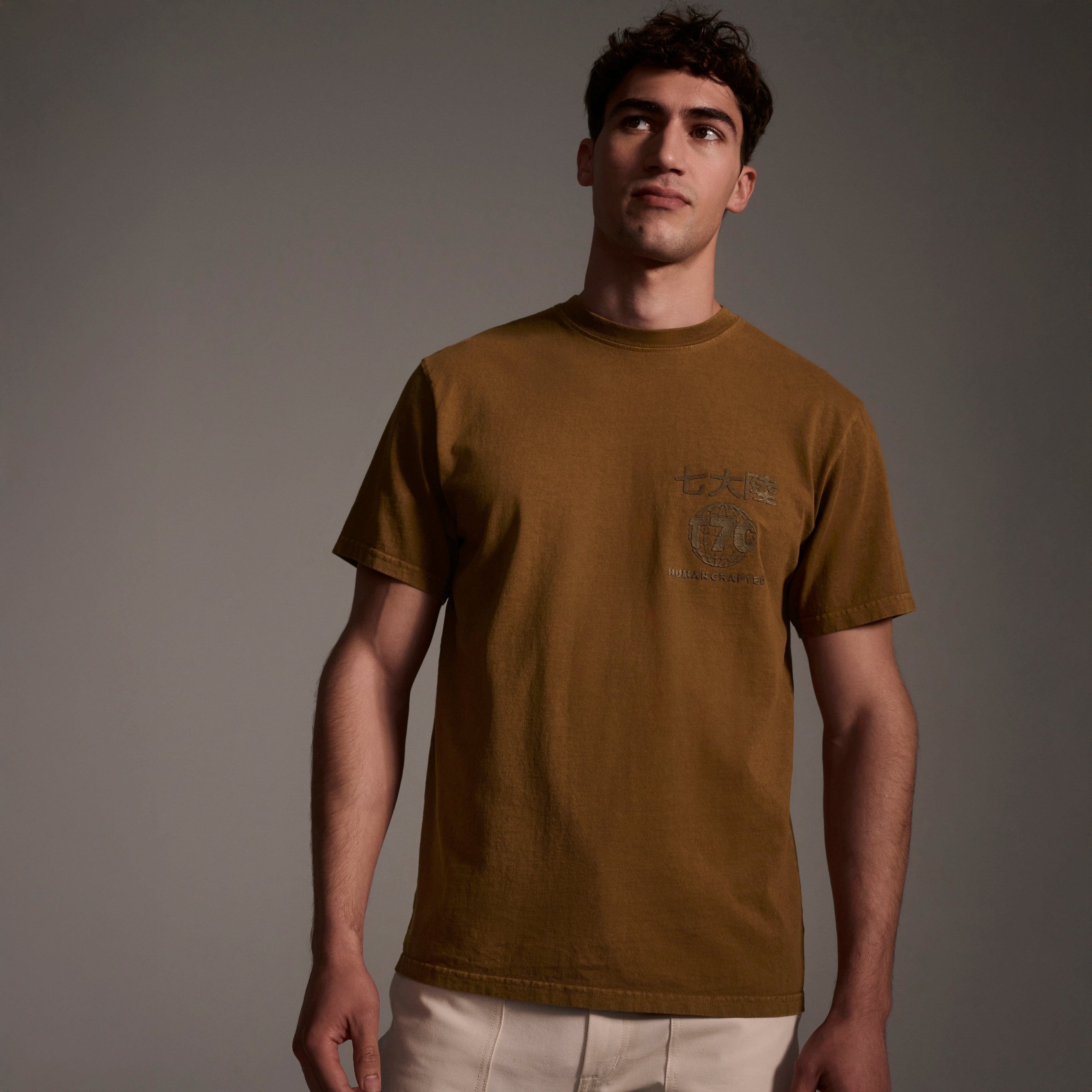 A young man models the Timo Tee in Tobacco by T7C The Seven Continents, paired with light-colored pants, standing against a softly lit gray background and gazing slightly upward.